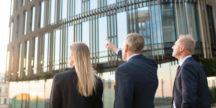 Group of business partners in formal suits pointing at office building, meeting outdoors, discussing real property. Back view. Commercial real estate concept