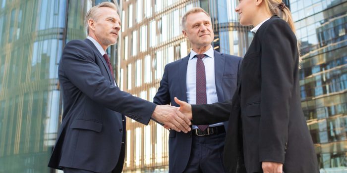 Positive business colleagues meeting in city, standing outdoors and shaking hands near office building. Low angle shot. Agreement and partnership concept
